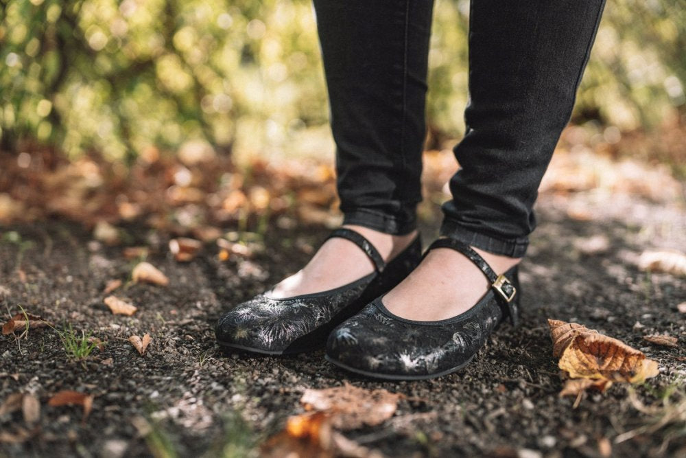 Nahaufnahme einer Person, die ZAQQs TWIST Ennis, schwarze Mary Jane-Schuhe mit Blumenmuster, und dunkle Jeans trägt. Sie steht auf einem Feldweg, der mit Herbstlaub übersät ist, während das verschwommene Grün im Hintergrund einen Hauch von heiterer Schönheit verleiht.