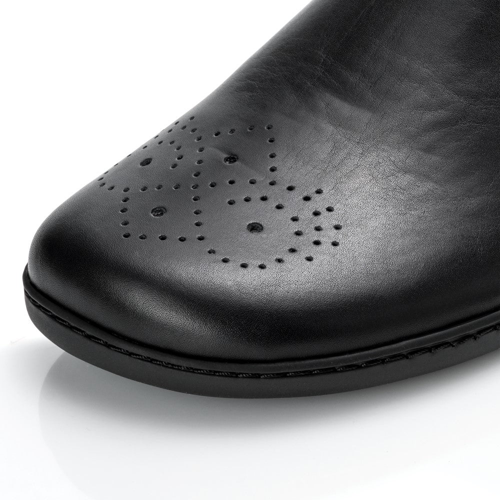 Nahaufnahme eines schwarzen ZAQQ EQUITY Brogue-Schuhs aus hochwertigem Nappaleder mit dekorativen Perforationen an der Spitze, die komplizierte Muster zeigen. Das elegante Chelsea-Boot-Design wird durch seine Platzierung auf einer reflektierenden weißen Oberfläche betont und unterstreicht die außergewöhnliche Handwerkskunst.