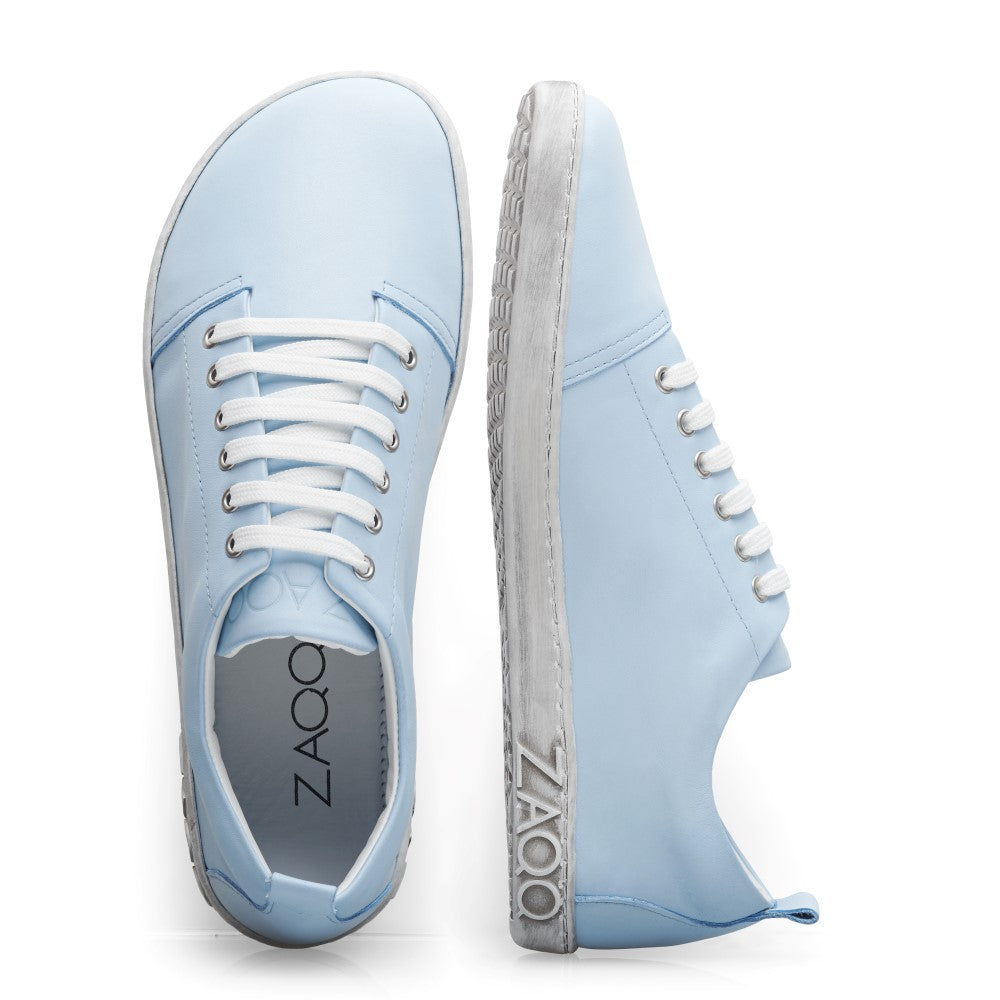 Die TAQQ Nappa Lightblue Sneaker von ZAQQ sind aus Premium Nappaleder gefertigt und verfügen über weiße Sohlen und Schnürsenkel. Ein Sneaker steht aufrecht und hebt die Schnürsenkel hervor, während der andere auf der Seite liegt und eine flexible Sohle mit dem geprägten ZAQQ-Markennamen zeigt.